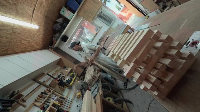 Dynamic wide angle view of a skilled woodcrafter evaluating a timber blank and selecting hand tools from a wall rack in a bright DIY garage workshop.
