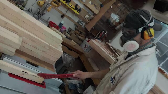 Unrecognizable male joiner wearing respirator mask and yellow earmuffs cutting timber block on table saw inside cluttered home workshop dynamic panning camera movement revealing authentic workspace