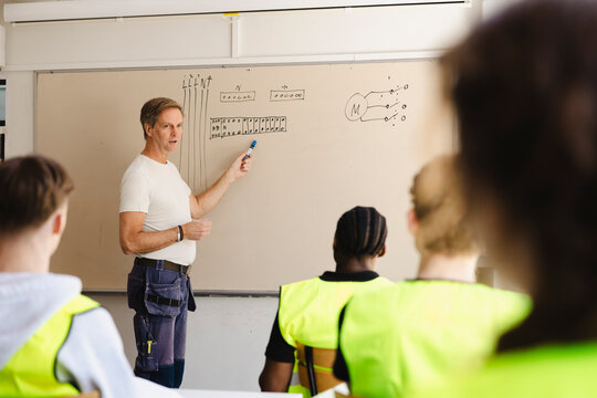 Male professor pointing at diagrams on whiteboard while explaining technical circuitry concepts to vocational students i
