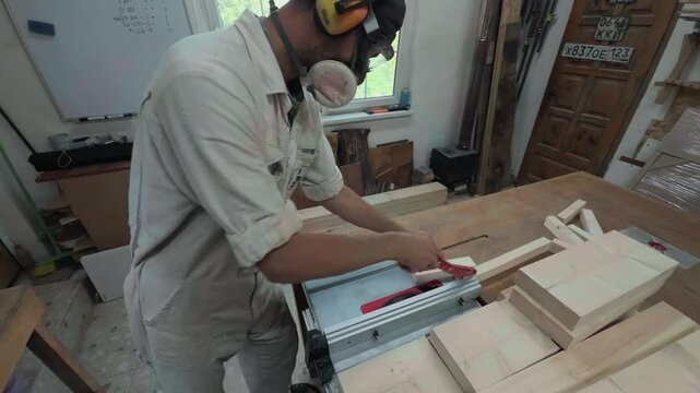 Unrecognizable male tradesman in hearing protection safely sawing a wooden batten with a red pusher on heavy machinery inside a rustic craft room dynamic sweeping arc shot of active labor
