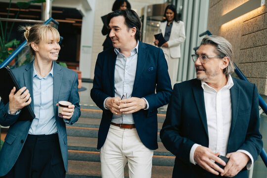 Smiling businesswoman talking with male colleagues while moving down on stairs in office building