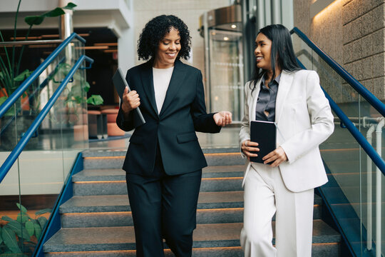 Smiling female business professionals talking with each other while moving down on stairs in office building