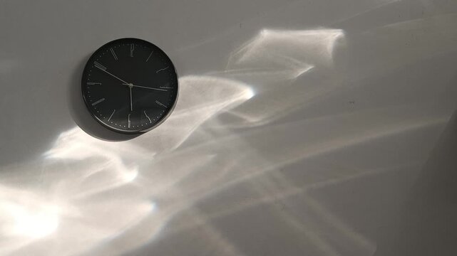Modern circular  gray or black clock against a white wall with sun reflections. Capturing the passage of time. 4K horizontal video.