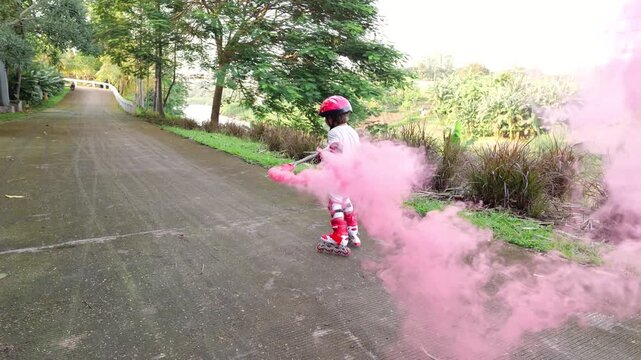 Young cheerful boy holding red smoke bomb on stick roller skating not very confidently along road, look back to trailing vibrant pink smoke. Slow motion shot, camera follow behind