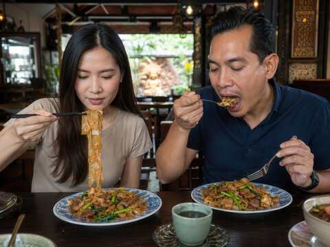 Junges Paar genie&szlig;t Pad See Ew mit breiten Reisnudeln, Rindfleisch und knackigem Gem&uuml;se in einem traditionellen thail&auml;ndischen Restaurant mit warmer, authentischer Atmosph&auml;re.