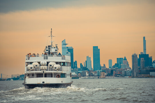 Passenger ferry leaving New York Harbor at dusk. Stern view shows a white vessel, upper deck, wake and distant Jersey City towers.