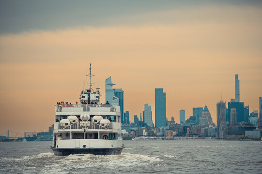 Passenger ferry on New York Harbor with skyline. Centered stern view of a white vessel moving across choppy water under an orange sky.
