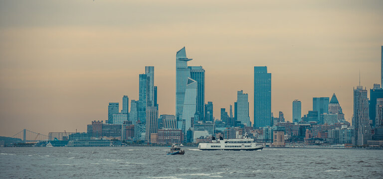 Manhattan skyline across Hudson River from a ferry. Panoramic frame shows towers, a passenger vessel and workboat on a gray channel.