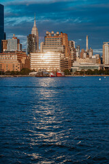 Fototapeta premium Waterfront buildings reflecting golden sunset light in Manhattan. Hudson River with ships docked at a pier and the Empire State Building behind.