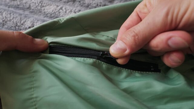 Human hands carefully fasten a black zipper on light green outerwear. The person slowly pulls the slider across the smooth fabric material. A textured gray surface sits behind.