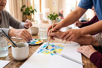 Obraz premium Elderly hands and caregiver hands work together on colorful watercolor painting at wooden table in warm care home room with cups, brushes, jars, and natural light for social creative activity