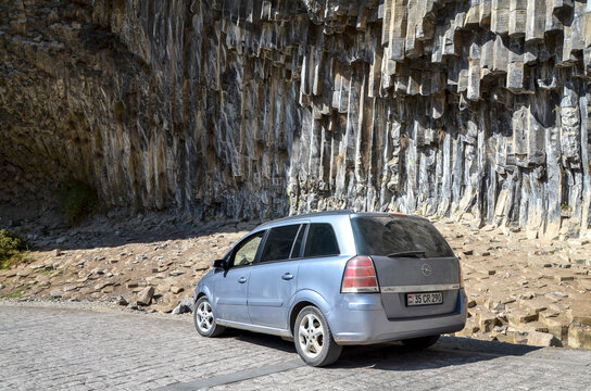 Grey family minivan Opel Zafira parked on a stone paved road in front of the Symphony of Stones basalt column cliff formation in Garni Gorge, Armenia 