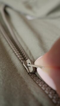 Fingers grip a metal zipper pull on a durable khaki green fabric garment in a macro view. The person slowly slides the slider along the plastic teeth to close the jacket or bag securely.