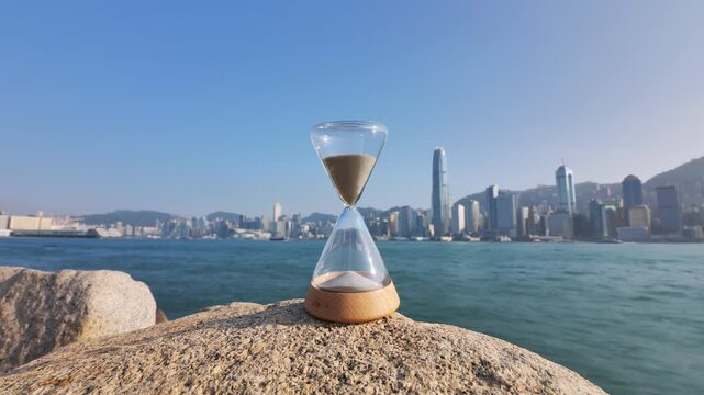 Time-lapse of hourglass standing at Hong Kong embankment with blurred Victoria Harbour and city waterfront on background. Conceptual shot of time's relentless passage with urban landscape backdrop