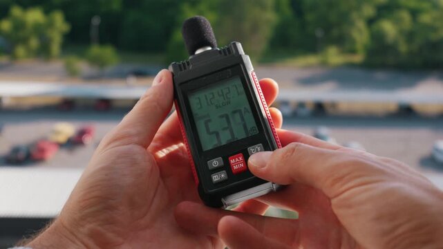 Close-up of hands holding a digital sound level meter displaying outdoor decibel measurements. Concept of noise pollution, environmental monitoring, and acoustic safety testing.
