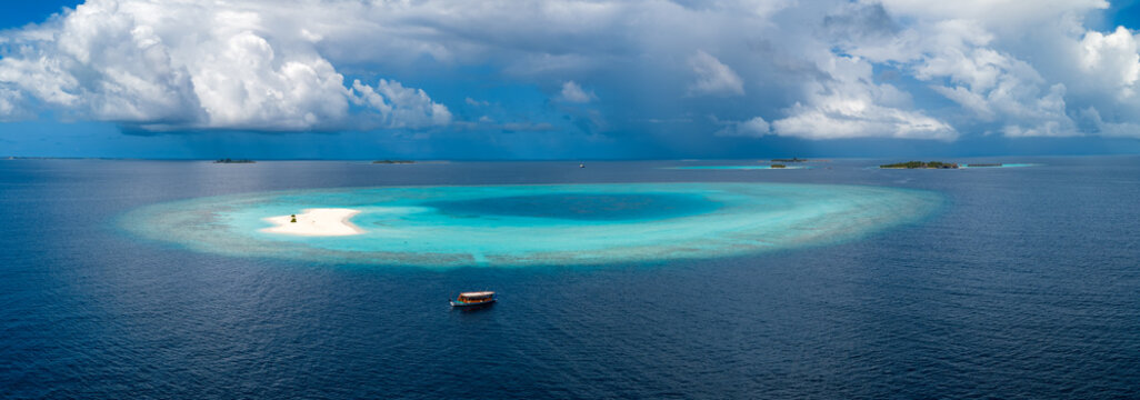 Aerial view of a circular sandbank and lagoon near Dhigurah, South Ari Atoll, Maldives. A dive boat drifts in deep sea, small vessels and islets sit under cumulus clouds.