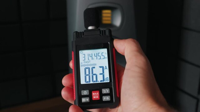 A hand holds a digital sound level meter displaying 83 decibels in a dark acoustic studio environment. Concept of sound engineering, noise pollution monitoring, and hearing protection.