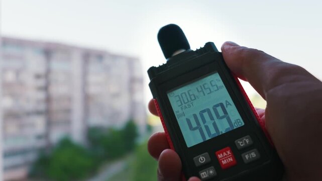 A hand holds a digital sound level meter displaying decibel readings against a blurred residential building. Concept of urban noise pollution and environmental acoustic testing.