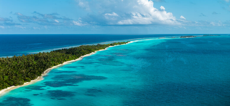 Aerial view of Dhigurah Island, Maldives, with elongated sandspit, reef edges, and lagoon. Turquoise shallows fade to deep cobalt ocean under midday light and clouds.