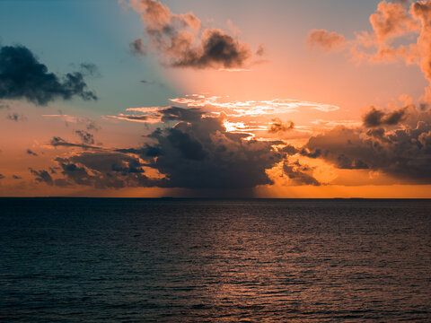 Sun sets near Dhigurah Island in the Maldives, with anvil clouds glowing orange and pink. Calm sea reflects teal and copper tones, suggesting a warm tropical dusk.