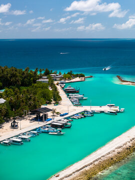 Aerial midday view of Dhigurah Island, Maldives, with slim sand and coral shore, compact marina, palms, narrow road, two boats leaving wakes toward the Indian Ocean.