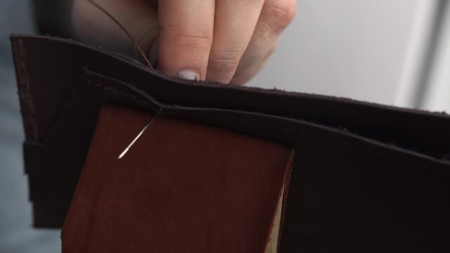 Leather craftsman hands stitching handmade brown wallet with needle and thread in a workshop, close up