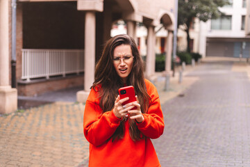 Obraz premium Distressed woman looking at her smartphone screen with an expression of shock and disbelief while standing outdoors in an urban environment wearing an orange sweatshirt and yellow-tinted eyeglasses