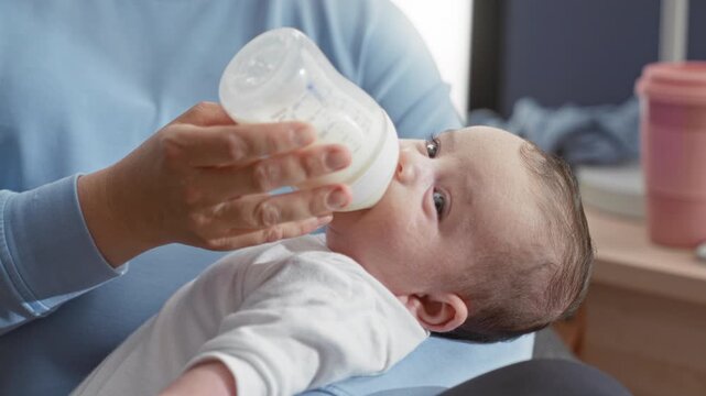 Newborn hispanic baby being bottle fed, eyes open, lying on caregiver lap in a home nursery beside a dresser and cup visible; comfort bonding.