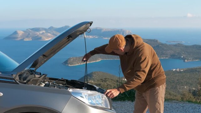 Man fixing broken down car with beautiful sea view. Young man trying to fix his broken down car with the hood open, stranded on a mountain road with a beautiful scenic sea view in the background