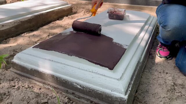 Woman applying protective paint coating with a roller on a concrete grave border during cemetery restoration and maintenance work in a European cemetery