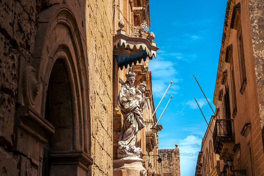 maltese limestone statue down alley narrow road in capital city valletta with clear blue skies overhead on sunny summers day next to brick building