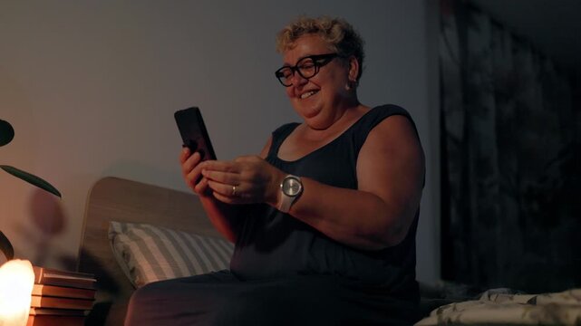 Overweight Happy Mature Woman Laughing With Smartphone In Bed At Night By Lamp Light