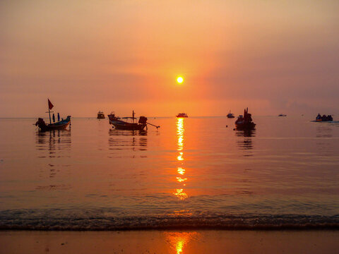 Hazy orange sunset with reflections over calm bay with boats in Thailand