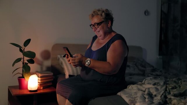 Overweight Relaxed Mature Woman Texting On Smartphone In Dark Bedroom With Lamp Light