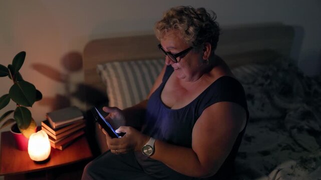 Overweight Cheerful Older Lady Watching Video On Smartphone In Bed At Night With Lamp