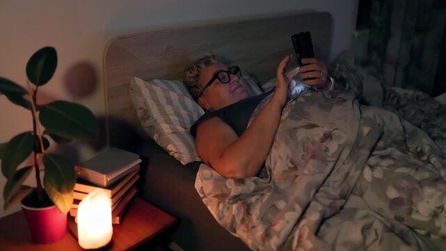 Overweight Mature Lady With Glasses Resting In Bed Browsing Smartphone At Night