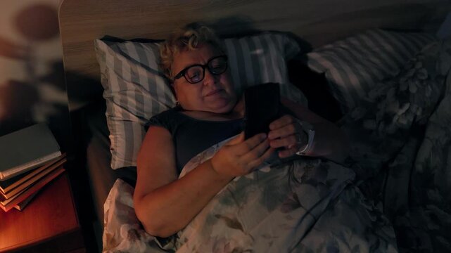 Overweight Senior Woman Smiling Viewing Social Media On Smartphone In Dark Bed