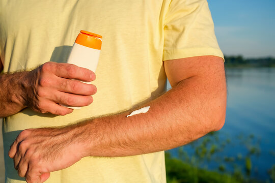 Man holding sunscreen bottle and applying cream to forearm outdoors by lake in summer