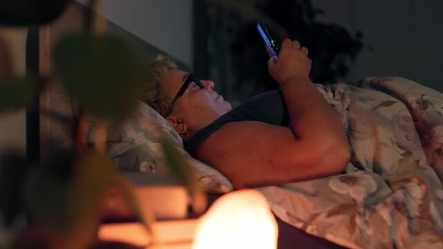 Overweight Joyful Senior Woman Texting On Smartphone In Dark Bed Illuminated By Screen
