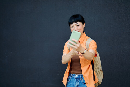 Portrait of a happy asian woman posing, using a smartphone phone or taking a selfie photo outdoors in front of a modern urban background