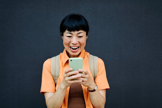 Portrait of a happy asian woman posing, using a smartphone phone or taking a selfie photo outdoors in front of a modern urban background