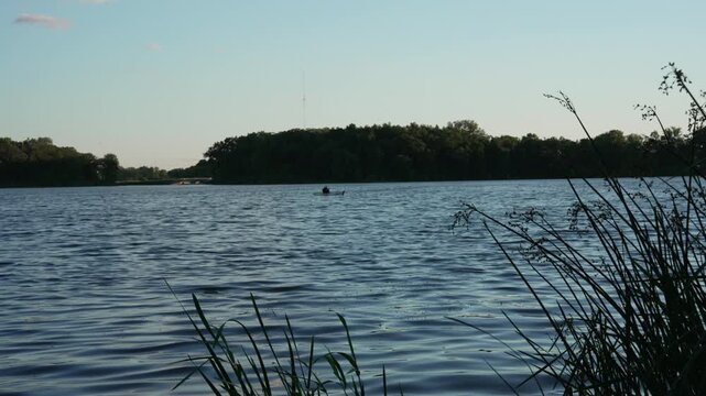 Quiet lake with gentle rippling water under a clear sky. In the distance, a lone person sits calmly in a kayak, enjoying the tranquility of nature. Outdoor recreation, summer relaxation, travel vlogs