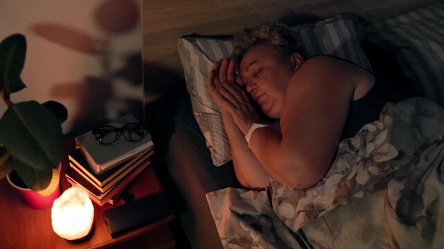 Overweight Mature Woman Sleeping Peacefully in Dark Bedroom Near Warm Lamp