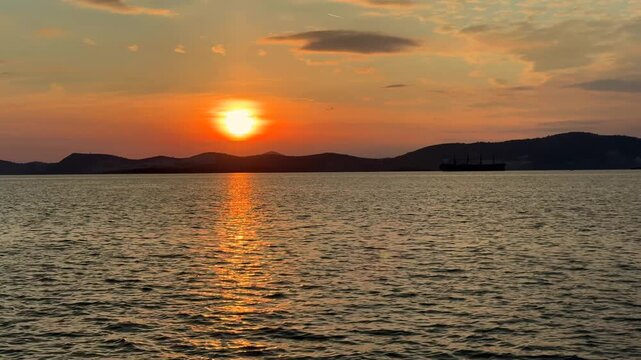 A calm, warm sunset over the Aegean Sea in Gulluk, Turkey. The sun hangs low in a hazy orange sky, casting a bright golden light path across the gently rippling water toward distant dark hills.
