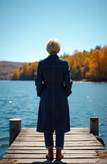 Fototapeta premium Mature woman stands on wooden pier looking at calm blue lake. Autumn trees with orange leaves surround the water. Clear sky above. Serene nature scene.