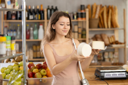 Surrounded by warm, homely ambiance of small grocery shop, young woman holding up packages artisan cheeses, comparing and contemplating choice with focused smile
