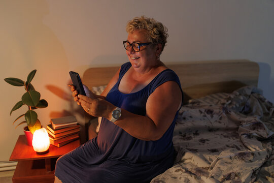 Happy overweight senior lady with glasses using mobile phone in dark bedroom.
