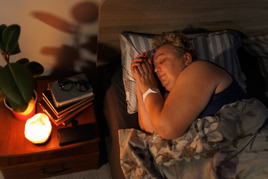 Senior overweight female sleeping in cozy bed near warm lamp and mobile phone.