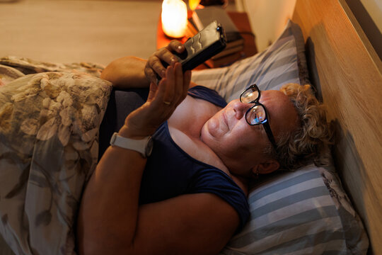 Overweight senior female using mobile phone with glasses resting in dark bed.