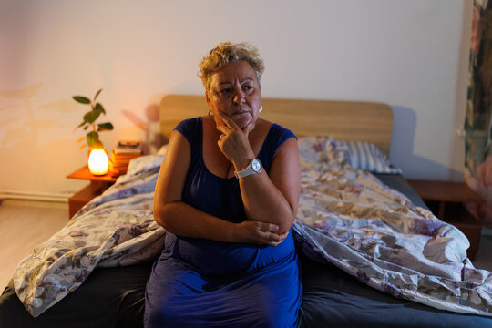 Sad overweight mature woman feeling lonely sitting in bed during dark night.
